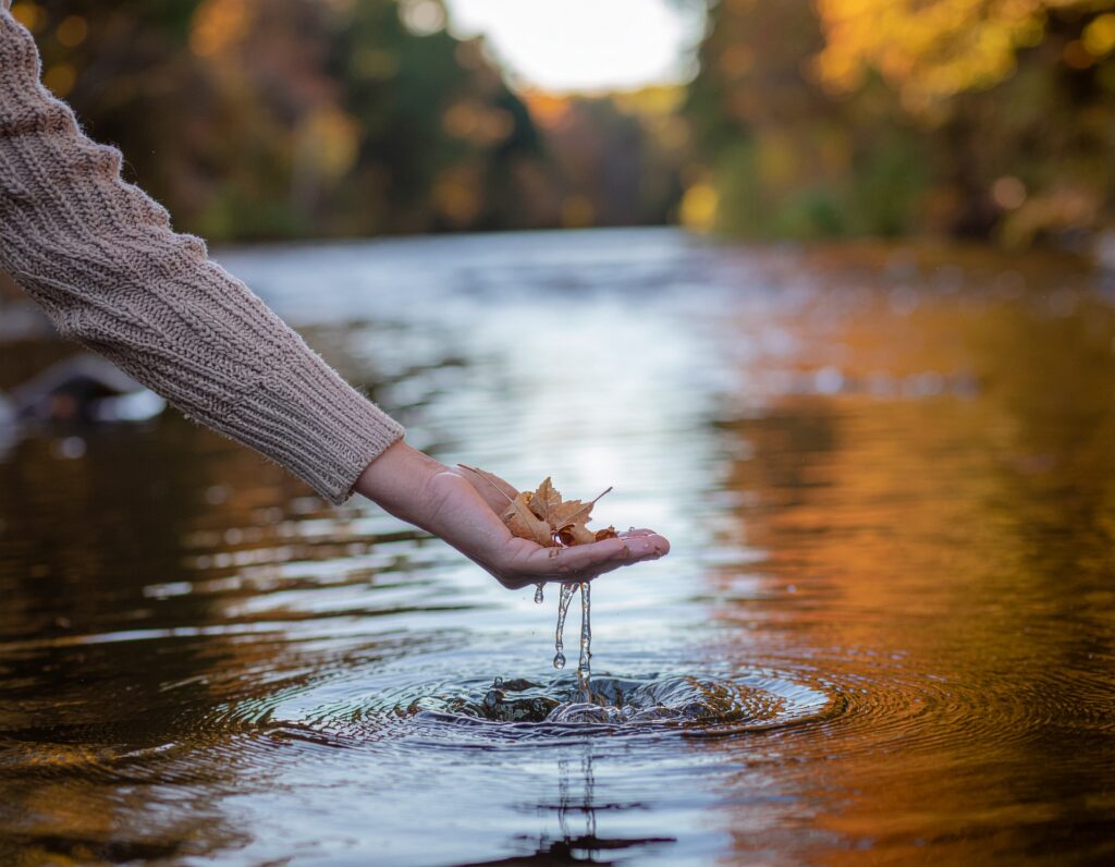 A close-up photograph of a hand emerging from a beige cable-knit sweater, extended over a clear autumn river. From the palm, water drips back into the water, creating concentric ripples that mirror the golden and brown tones of the fall foliage bokeh in the background.