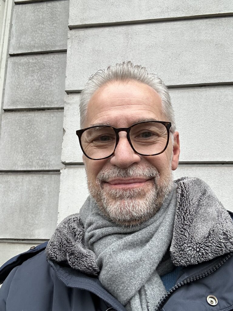 A natural-light portrait of author Jens Neitzel with grey hair and a white beard, smiling calmly and directly at the viewer. He wears a dark navy jacket over a grey top. The background is a softly blurred, serene view of a calm lake and distant hills under a gentle, cloudy sky, conveying a sense of tranquility.